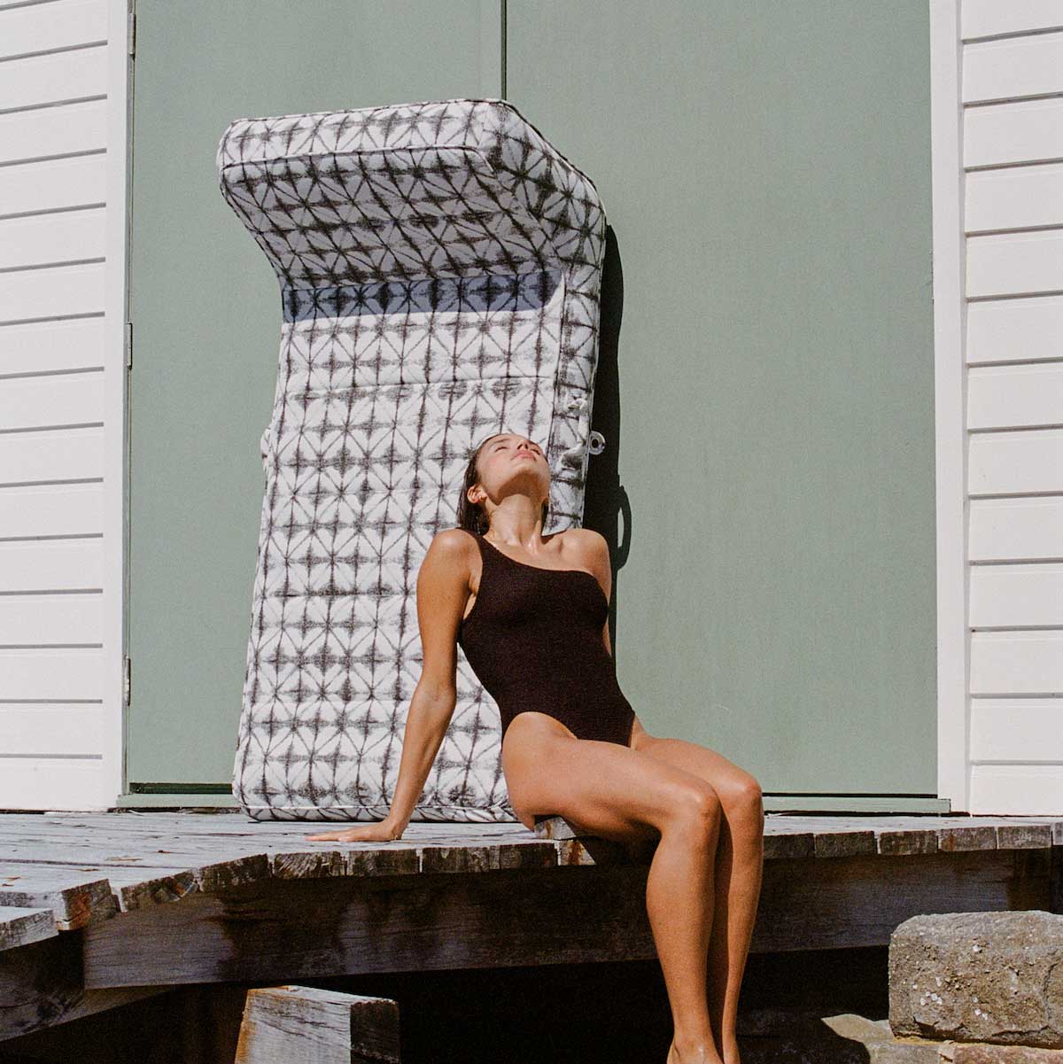 A women sitting on a wooden pier with a luxury pool floats for adults leaning against the wall behind her.
