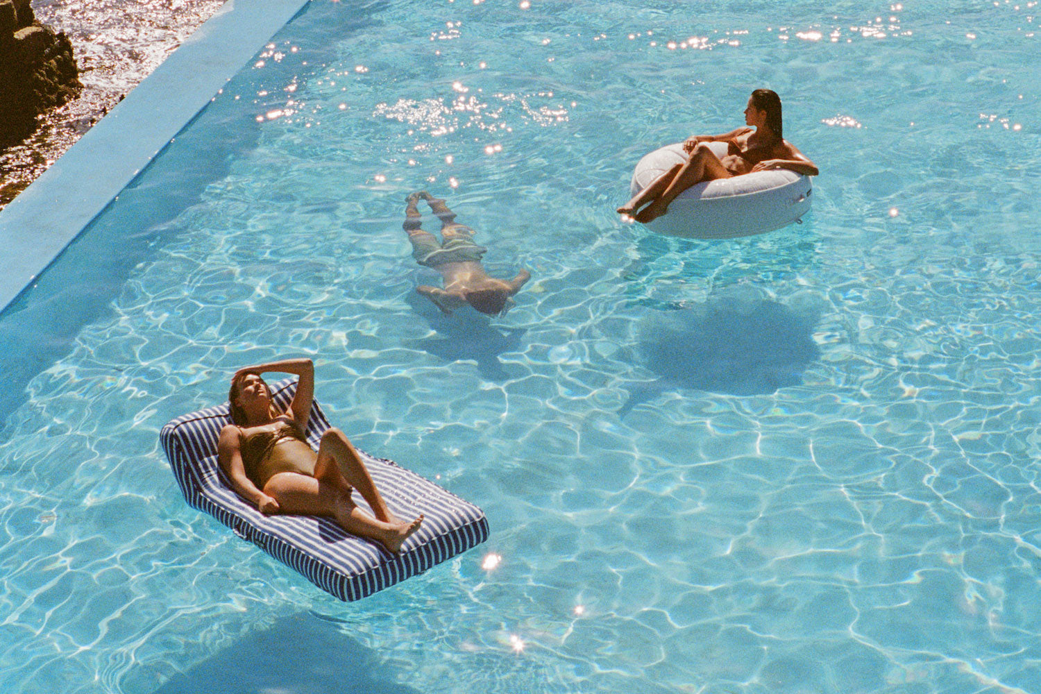 Two adults on pool floats for adults in a swimming pool with a man swimming beneath them underwater. 