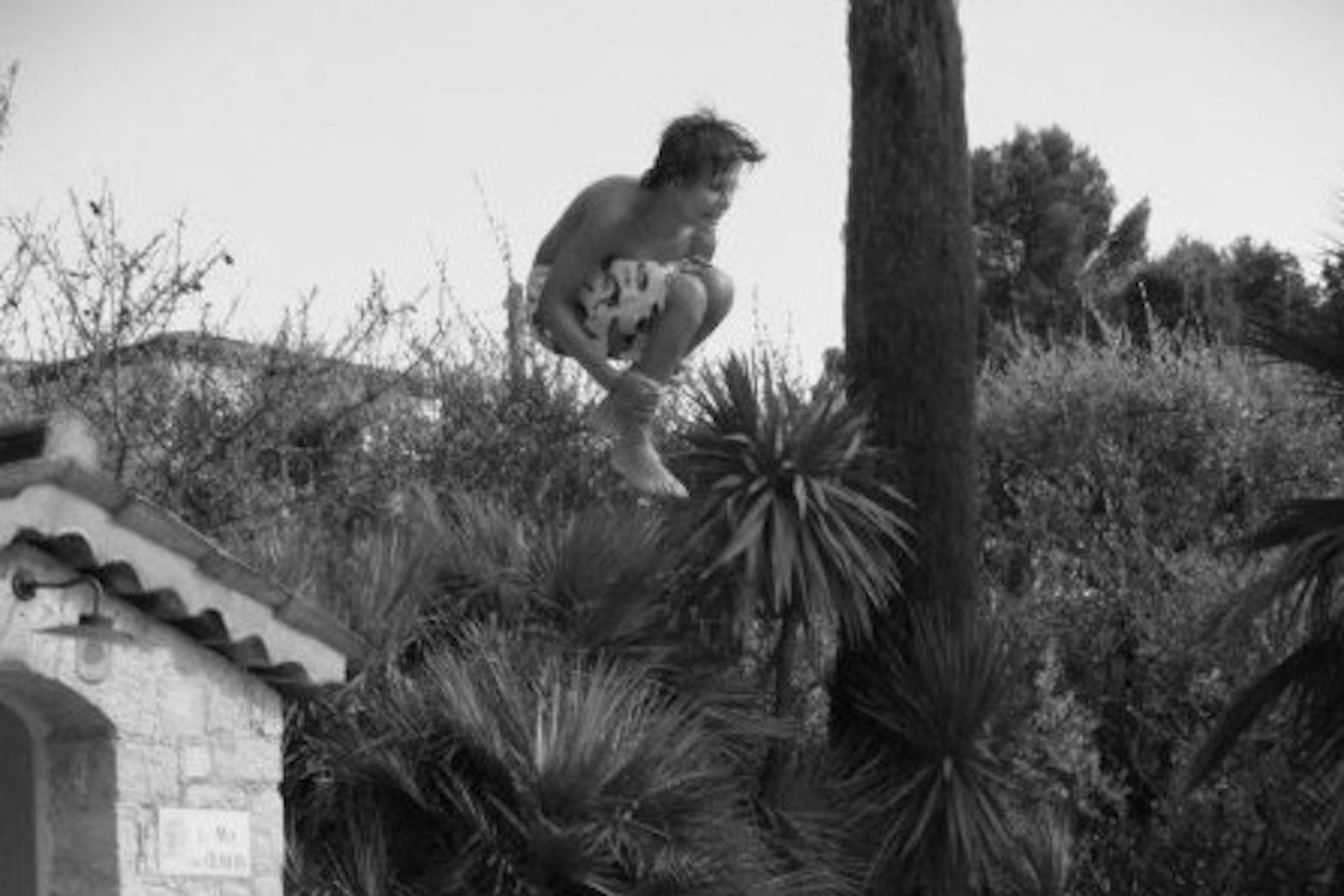 A boy jumping from the top of a pool house cannonballing into a swimming pool with trees and sky in the background.