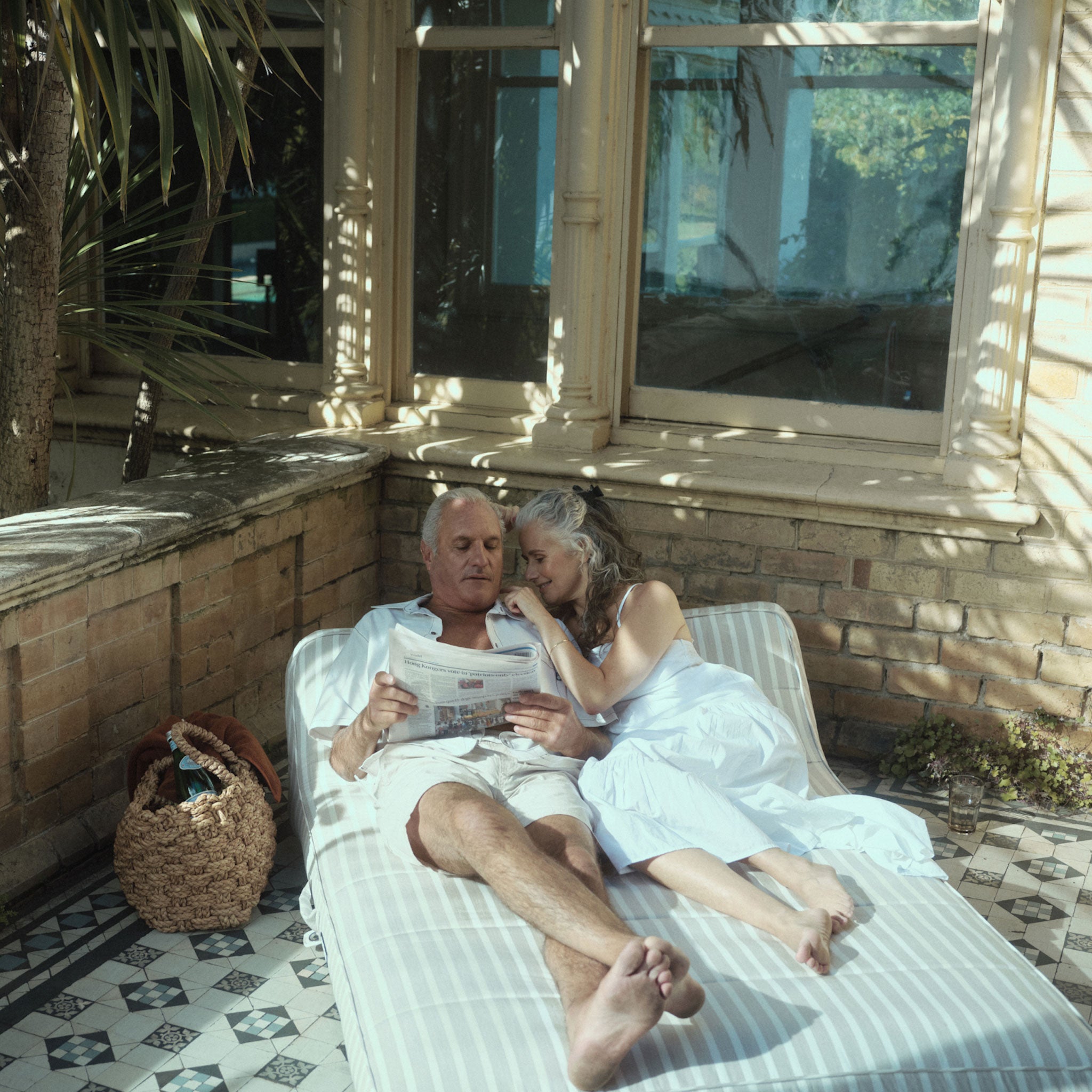 An elderly couple sitting on a beige and white striped pool float lounger for adults on the terrace of a home.