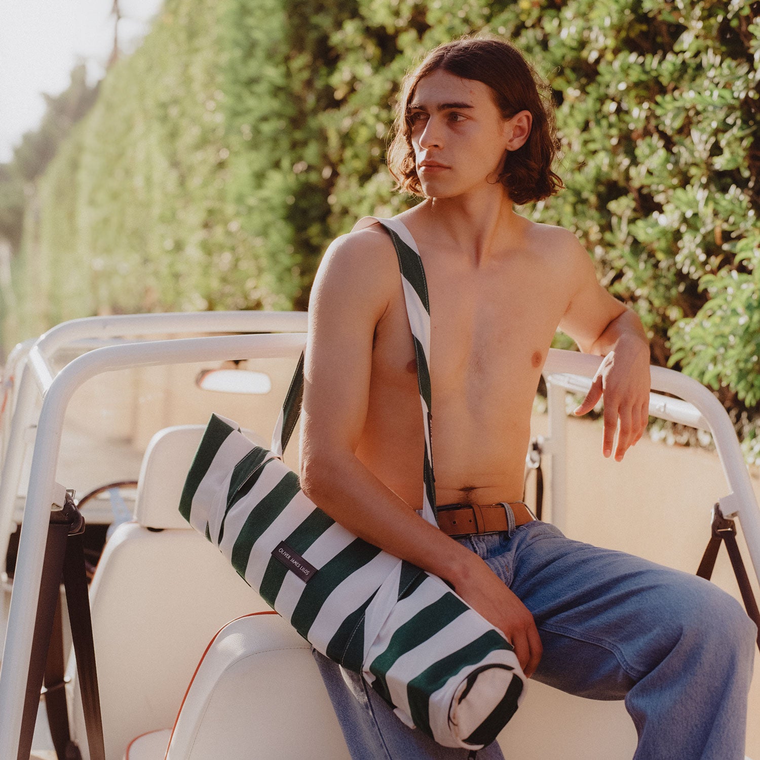 A man sitting on a white vintage car with a deflated pool hammock for adults around his shoulder.