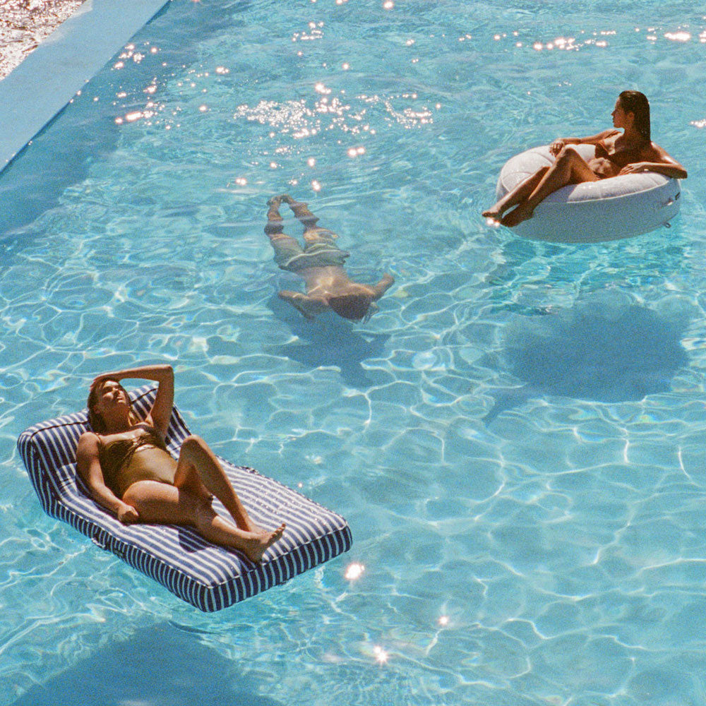 Two women lying on luxury pool floats floating in the water with a man swimming underwater.