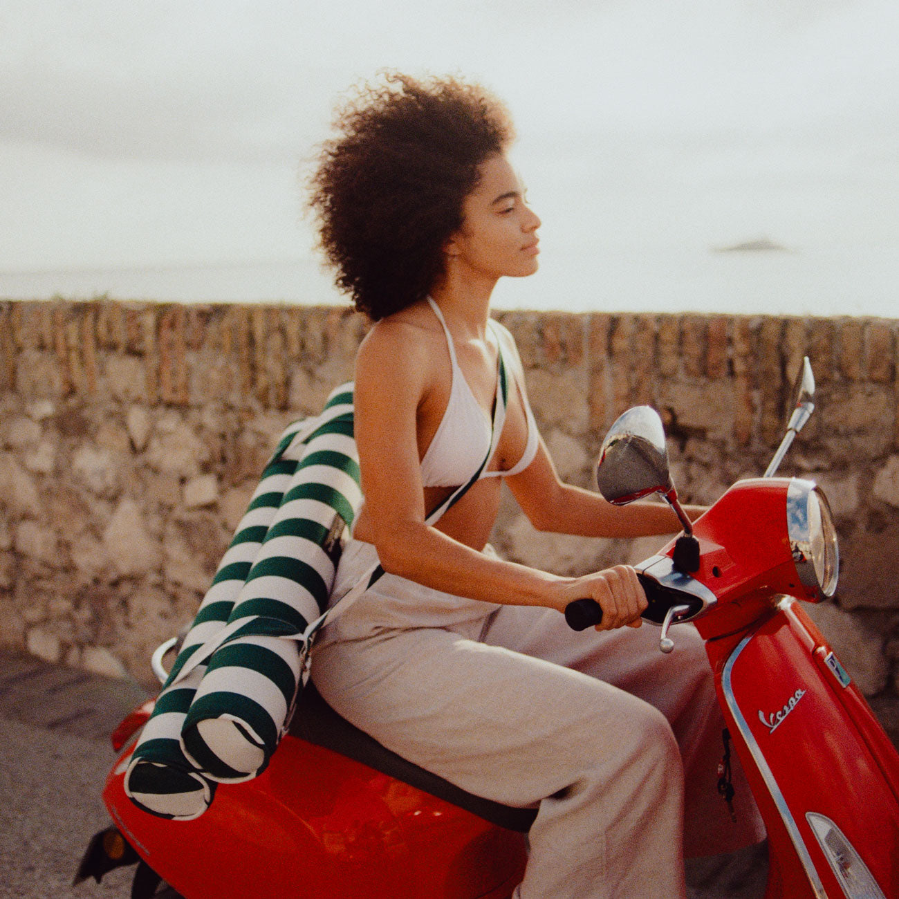 A women on a red moped driving next to the sea with a luxury beach float for adults strapped on her back.