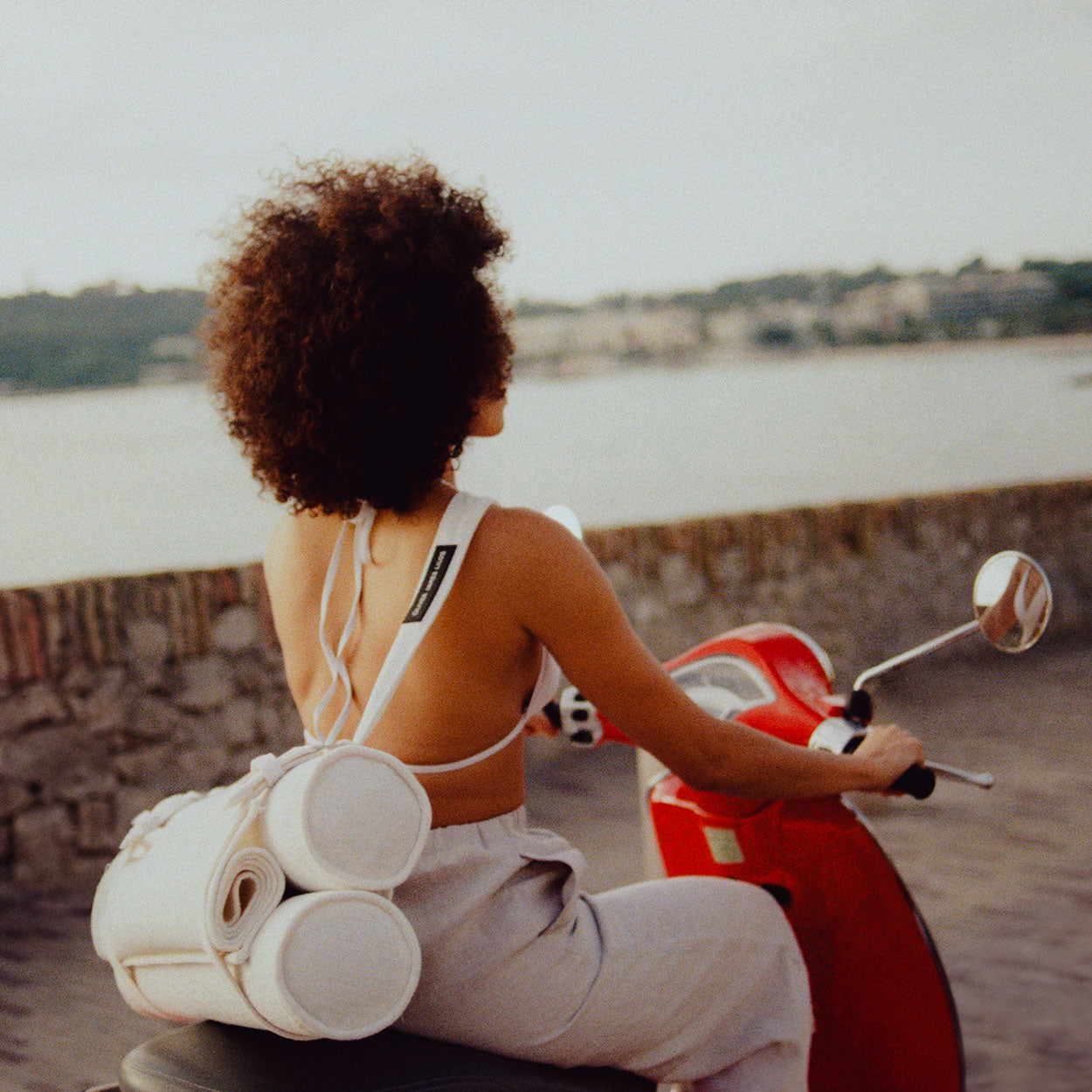 A women on a moped with the beach int he background and a beach float for adults strapped to her back.