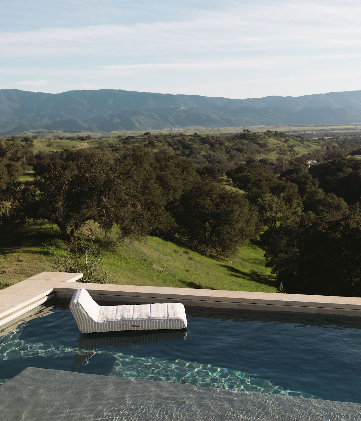A luxury beige and white stripe pool float lying in a swimming pool with a view of trees, hills and mountains.