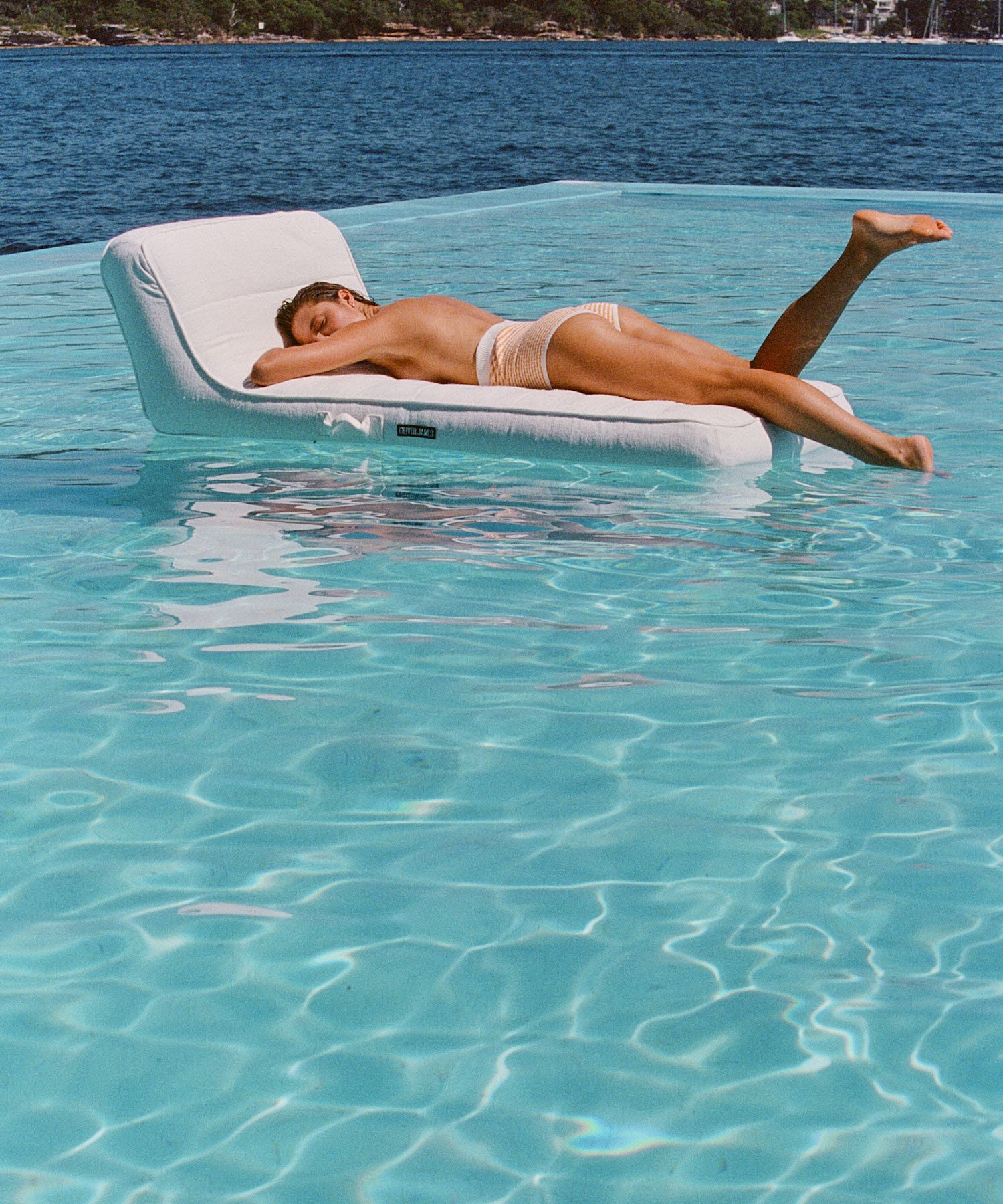 A women lying face down on a single luxury pool float lounger with her toes dipping into the water in a swimming pool overlooking the ocean.