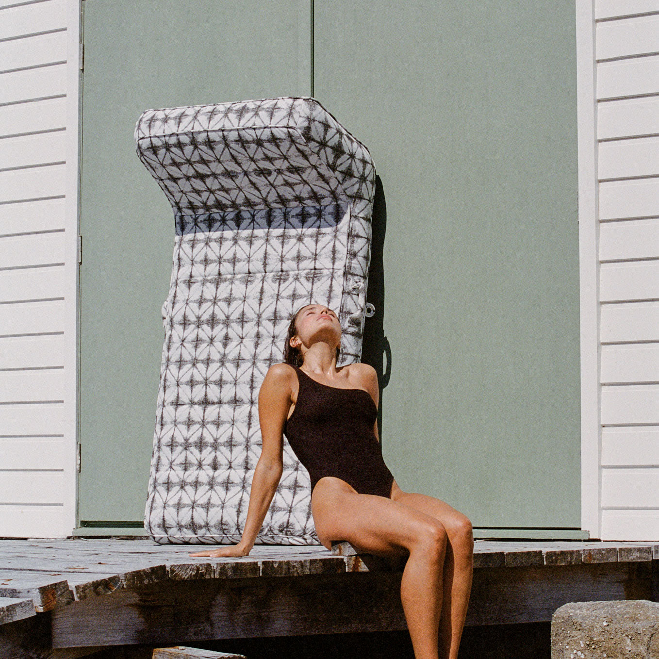 A women sitting on a platform with her head back with a white and grey luxury pool float lounger leaning against a pool house in the background.