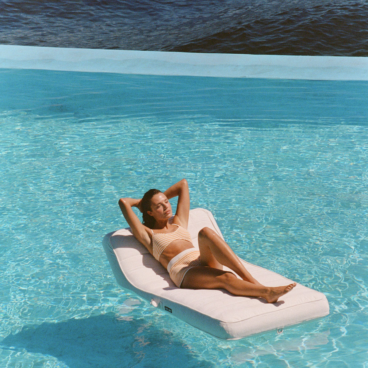 A women with her hands behind her head floating on a luxury pool float lounger in a swimming pool with the ocean in the background. 