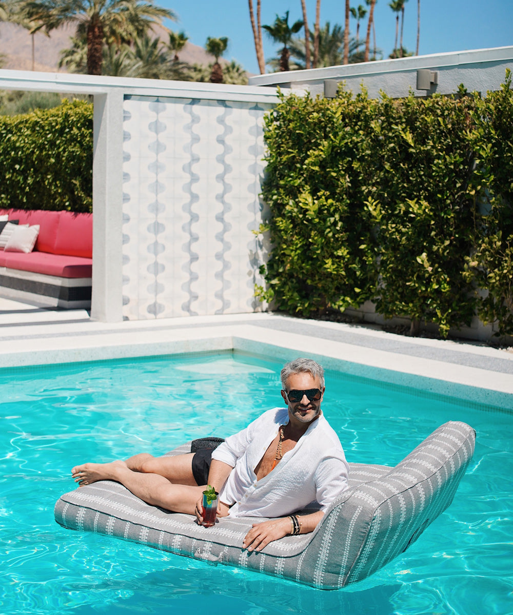 A man sitting on a grey and white striped luxury pool floats holding a drink.