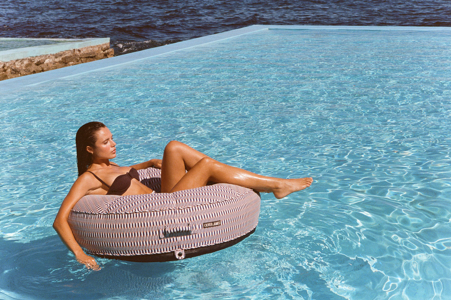 A woman sitting on a red and white luxury pool float in a pool with the ocean in the background.