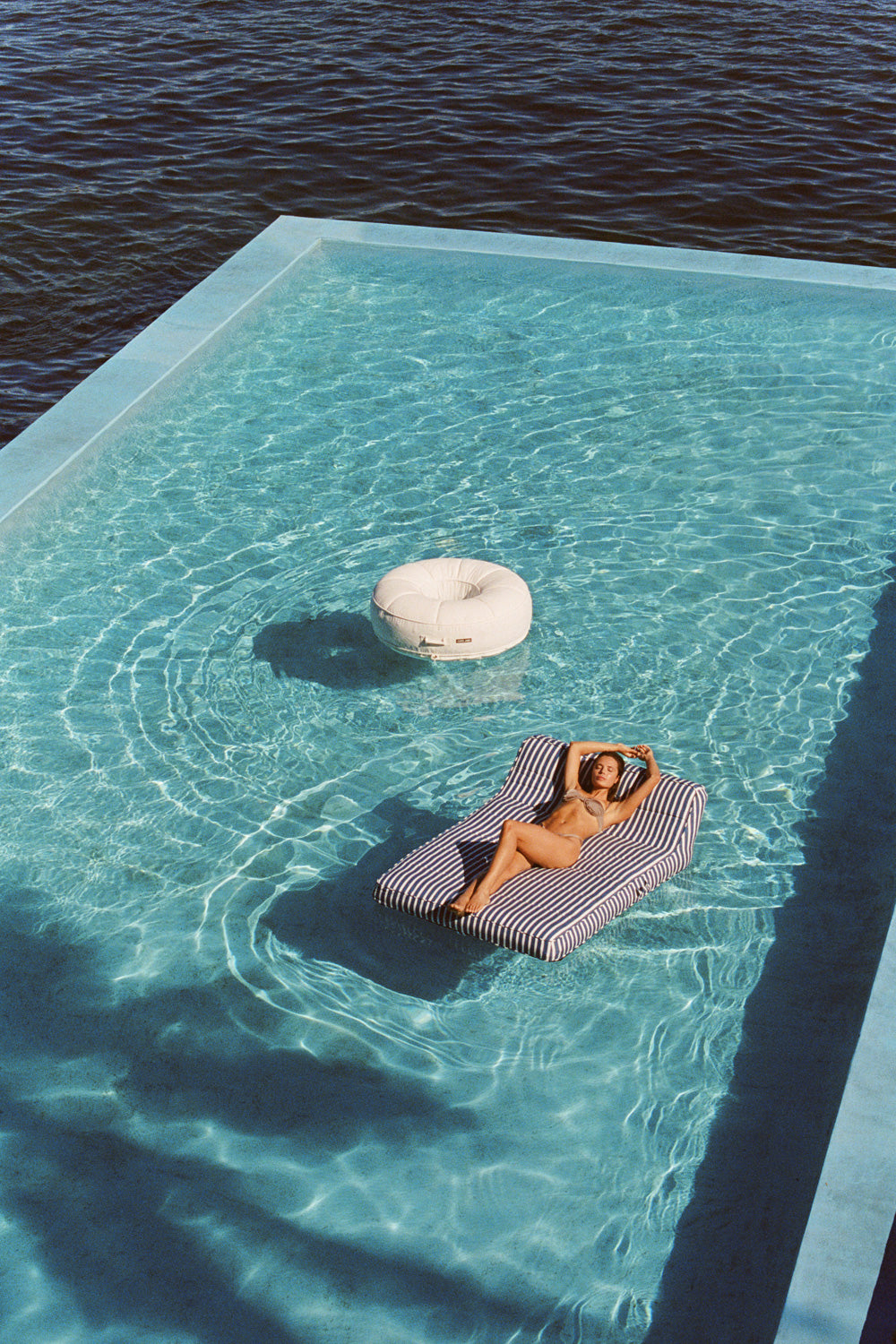 A women floating on a double blue and white striped luxury pool float in a pool with the ocean and white ring luxury pool float in the background.
