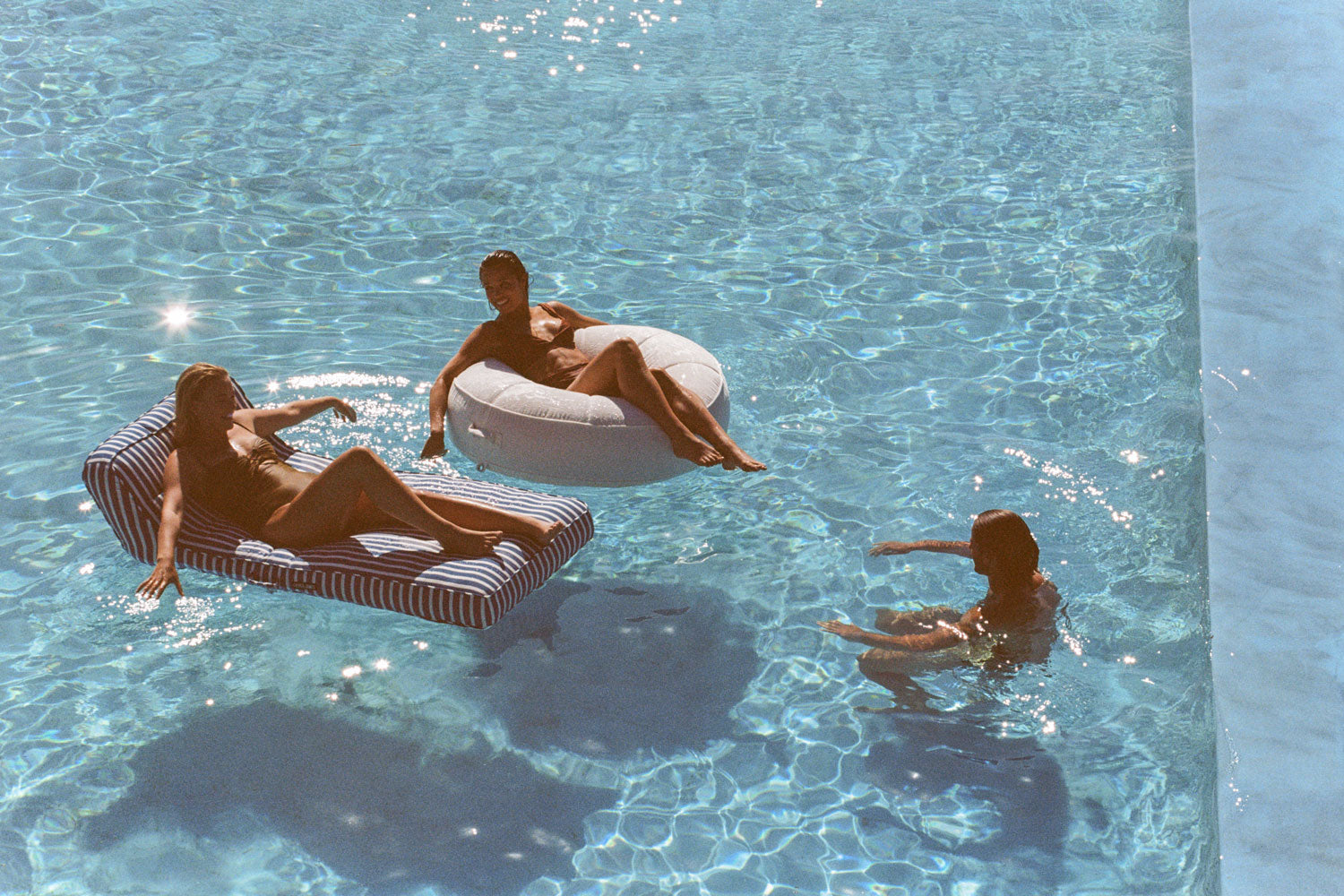 Two women lying on luxury pool floats in a swimming pool with a man swimming besides them.