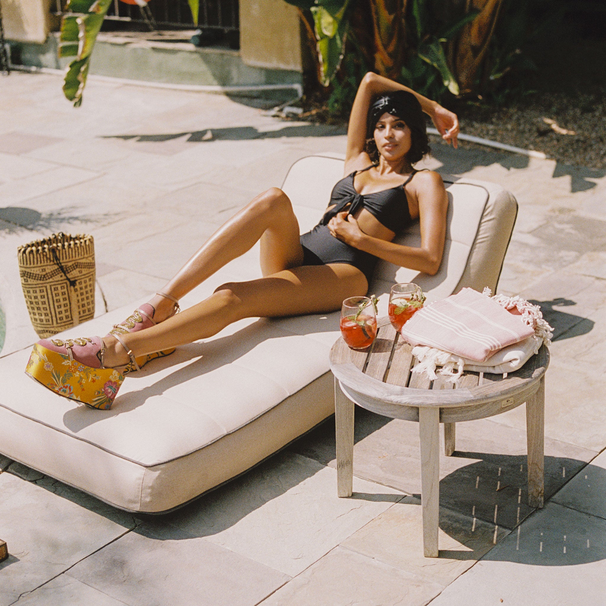 A women laying on a brown tan luxury pool float by the side of a swimming pool with a basket and table next to her.