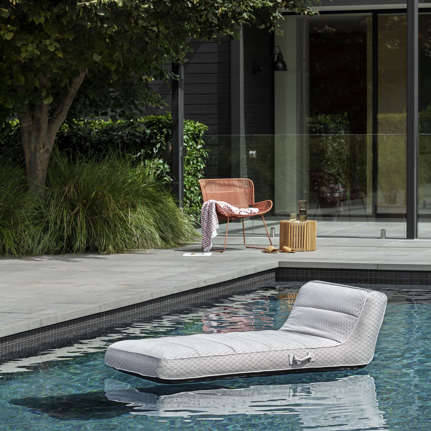 A black and silver luxury pool float floating in a swimming pool with a chair and glass pool house in the background.