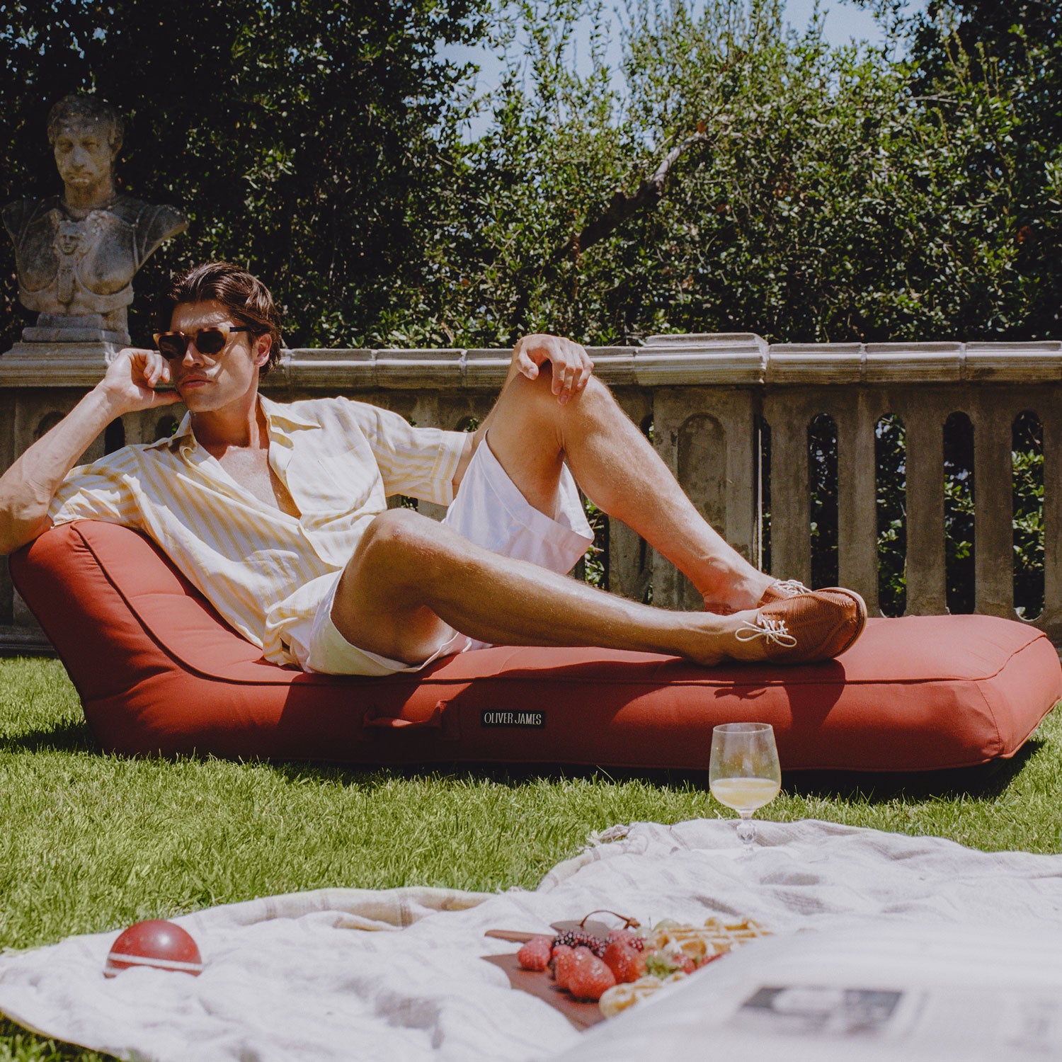 A man in sunglasses laying on a terracotta colored luxury pool float on a grass with a picnic.