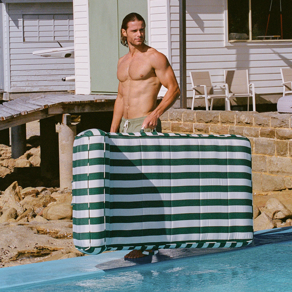 A man carrying a green and white striped luxury pool float on the edge of a swimming pool with a pool house and rocks int he background.