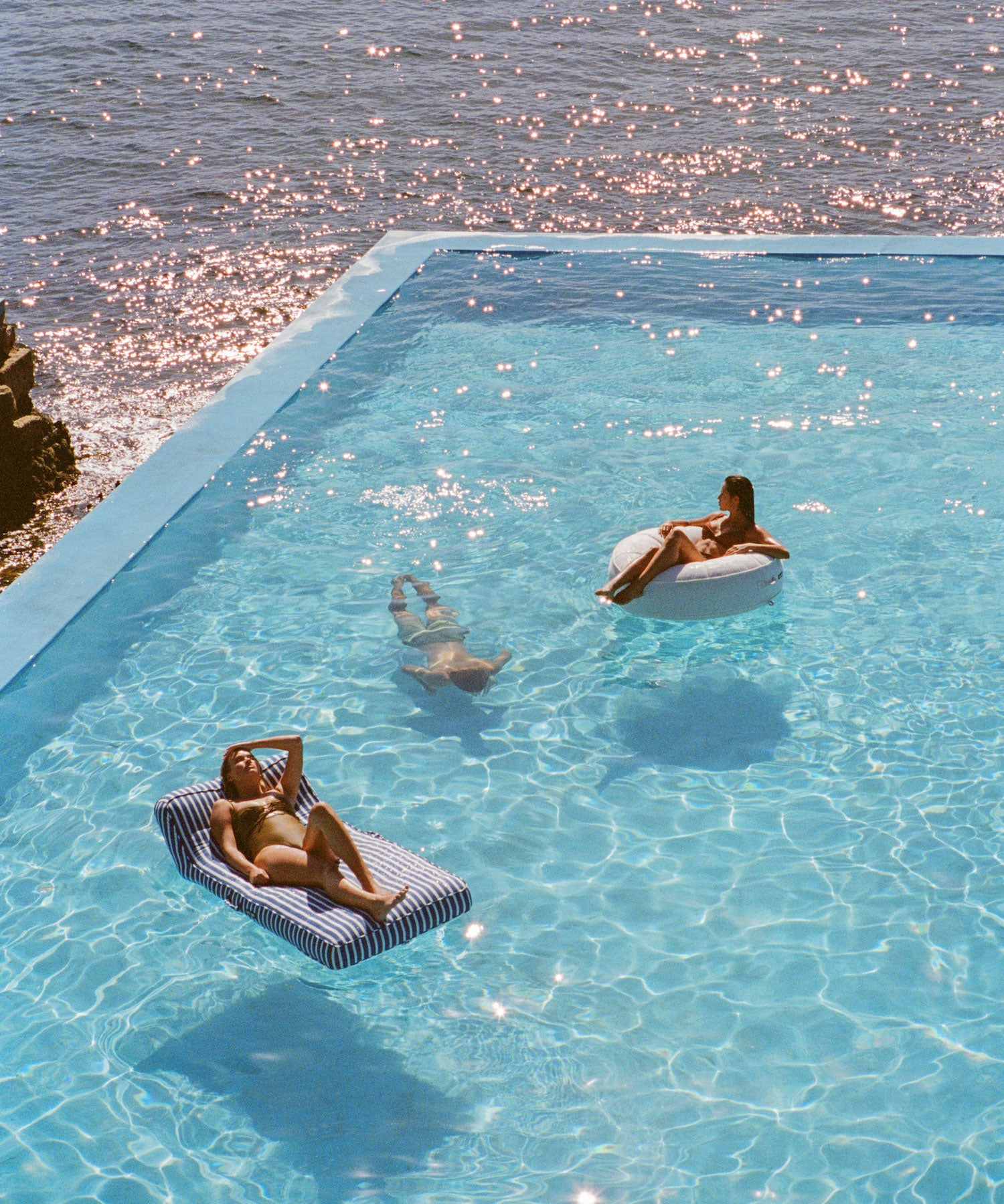 Deux adultes sur des flotteurs de piscine pour adultes dans une piscine surplombant l'océan avec un homme nageant en dessous d'eux sous l'eau. 