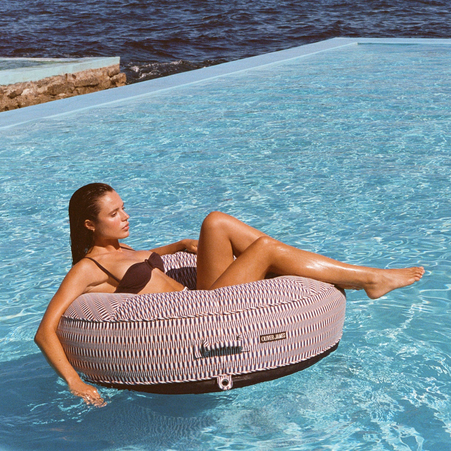 A women on a red, white and orange ring pool float lounger for adults floating in a swimming pool with the ocean in the background.