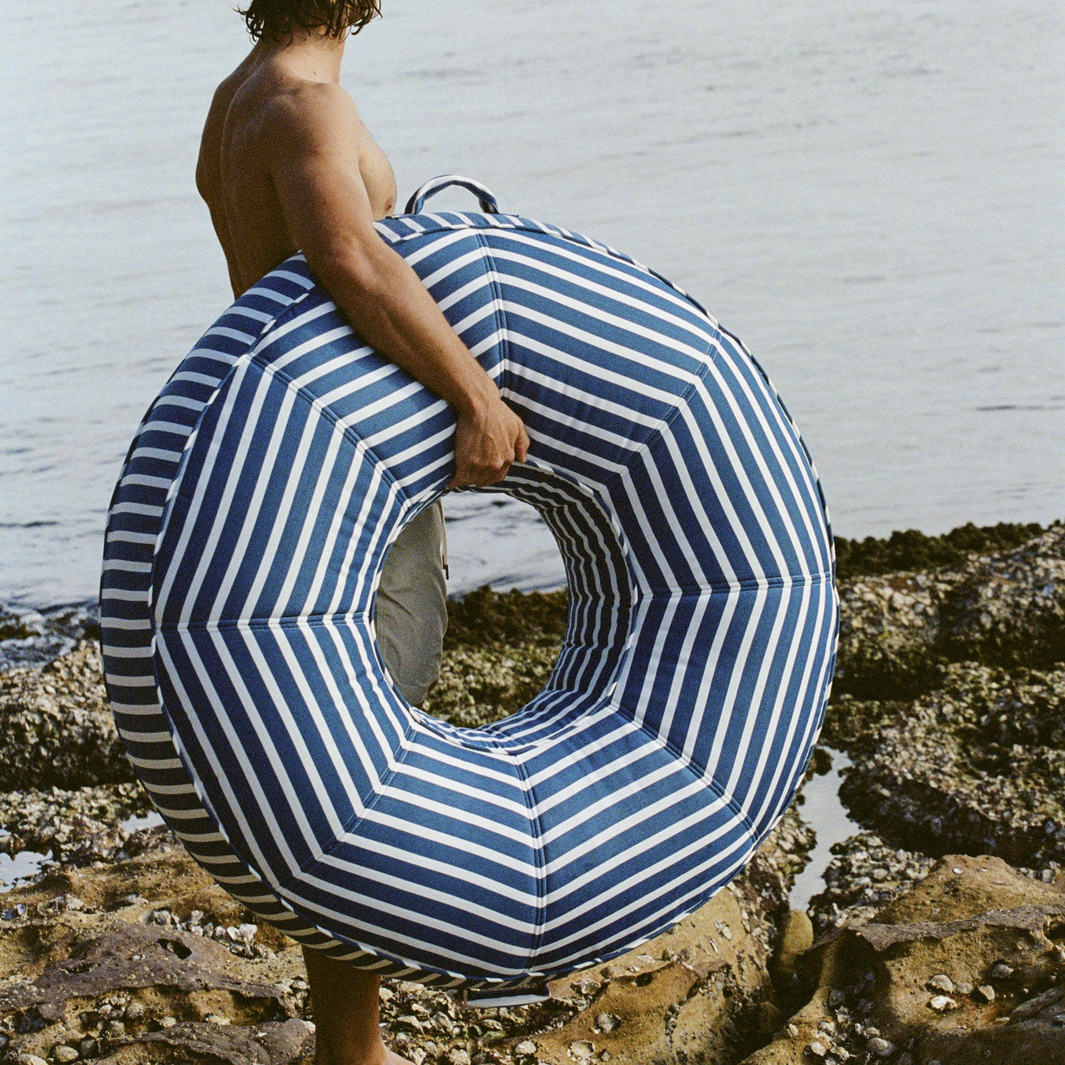 A man carry a blue and white striped fabric ring pool float lounger in one hand on a rocky beach near the ocean. 