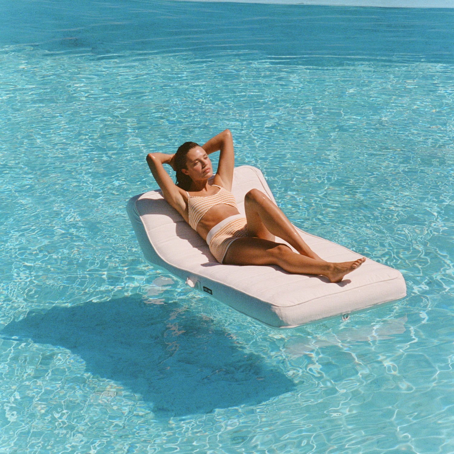 Une femme flottant sur un flotteur de piscine blanc pour adultes, les bras au-dessus de la tête, dans une piscine bleue.