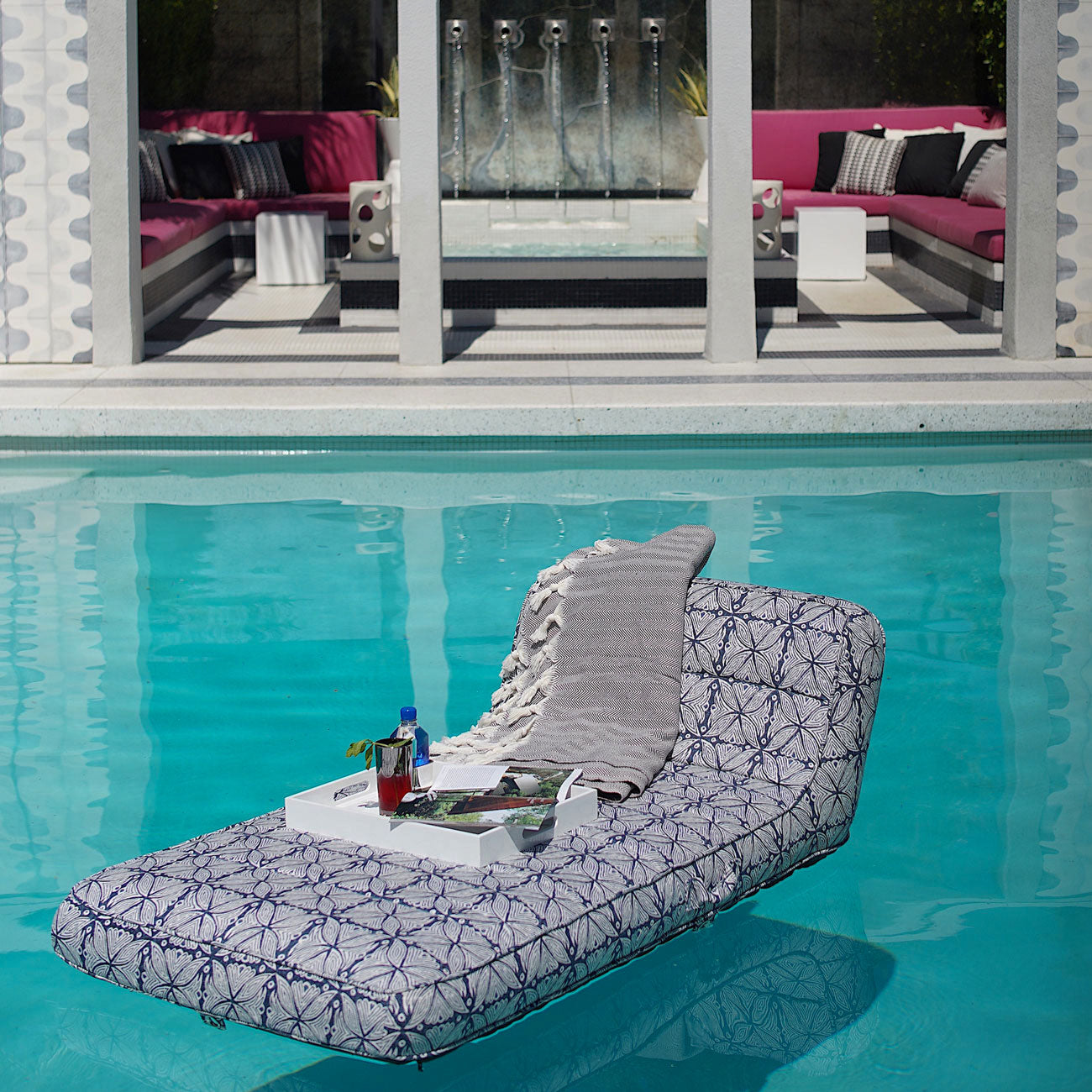 A blue and white patterned pool float lounger for adults floating in a swimming pool with a tray and throw over the lilo.