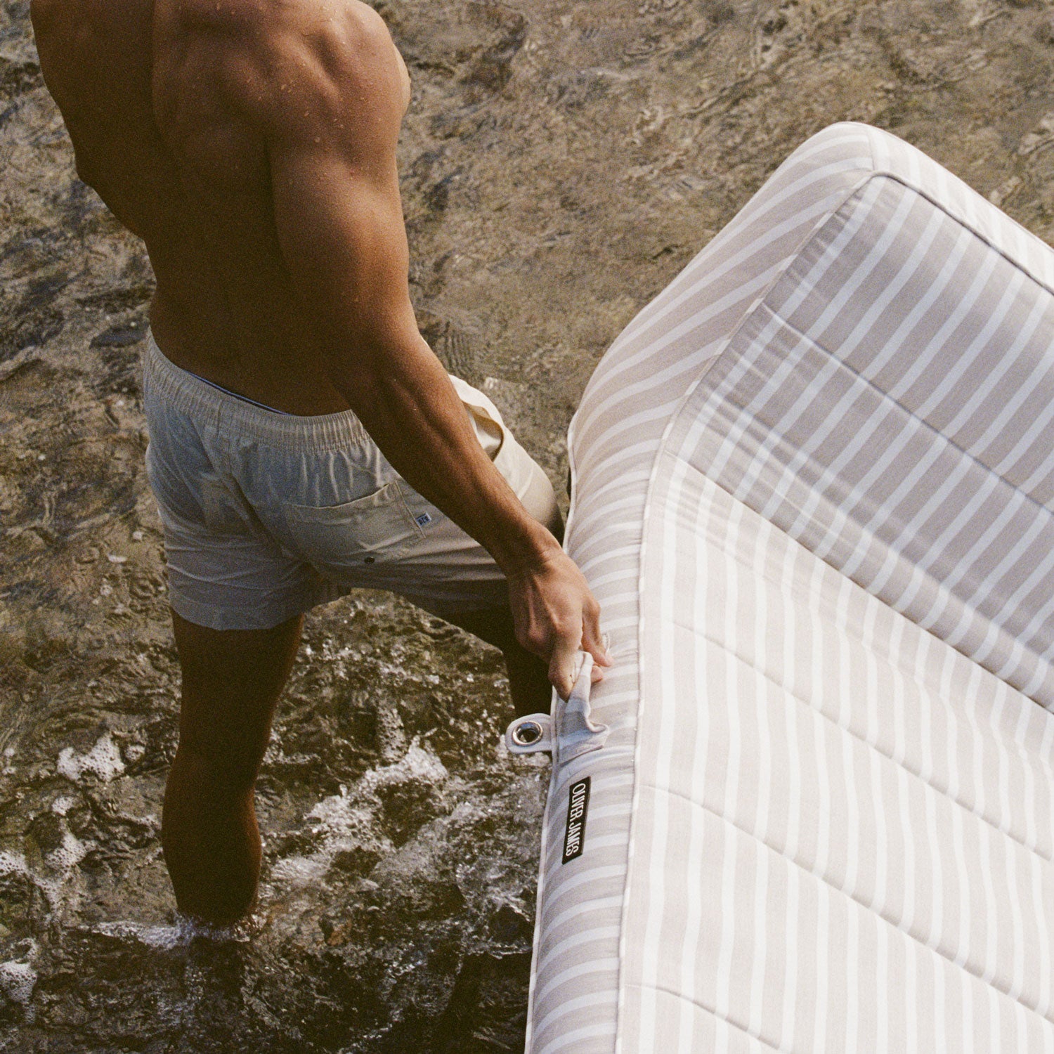 Shoulders down of man carrying a white and beige striped pool float lounger for adults over rocks at a beach.