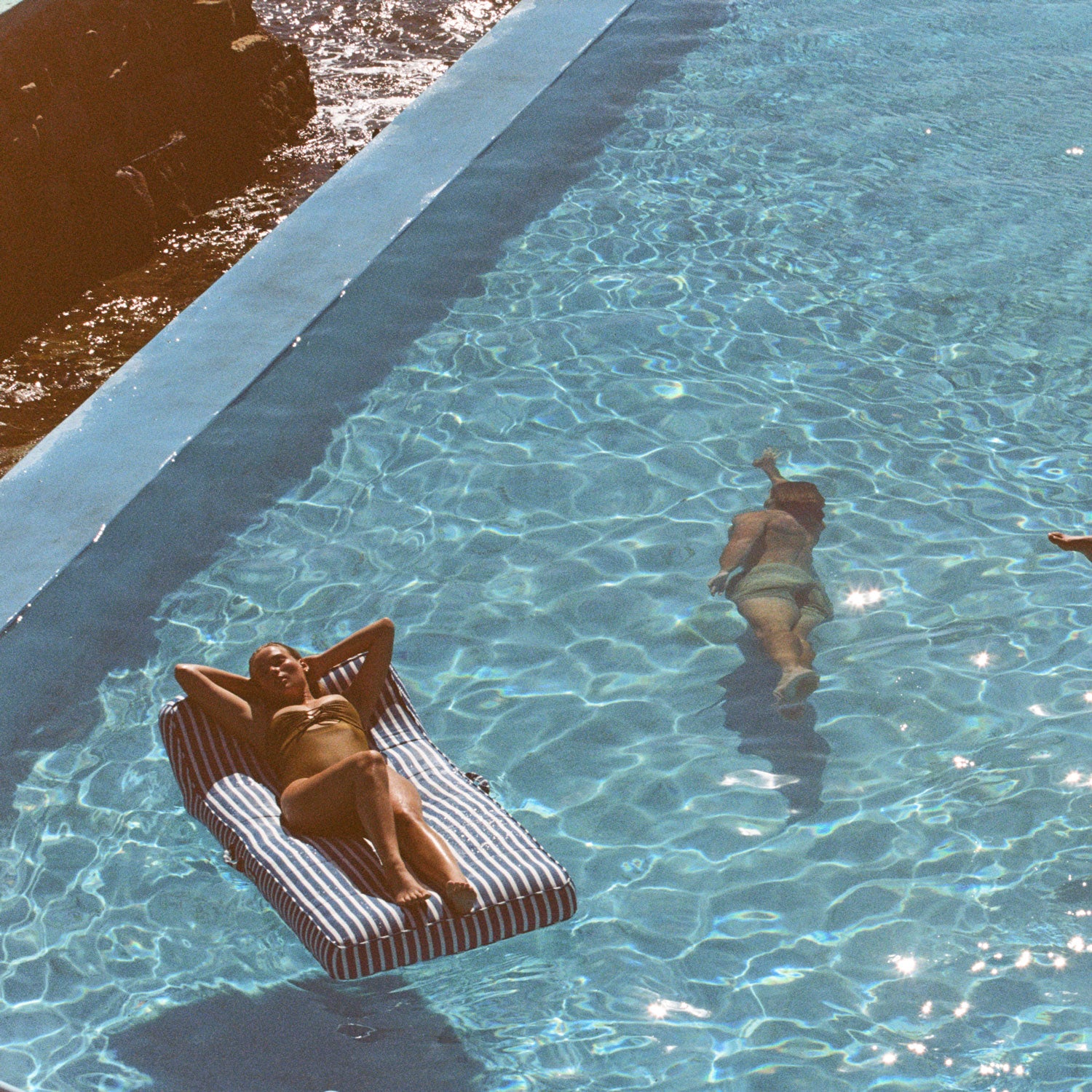 Une femme sur un flotteur de piscine bleu et blanc pour adultes flottant dans une piscine avec des rochers en arrière-plan et un homme nageant sous l'eau.