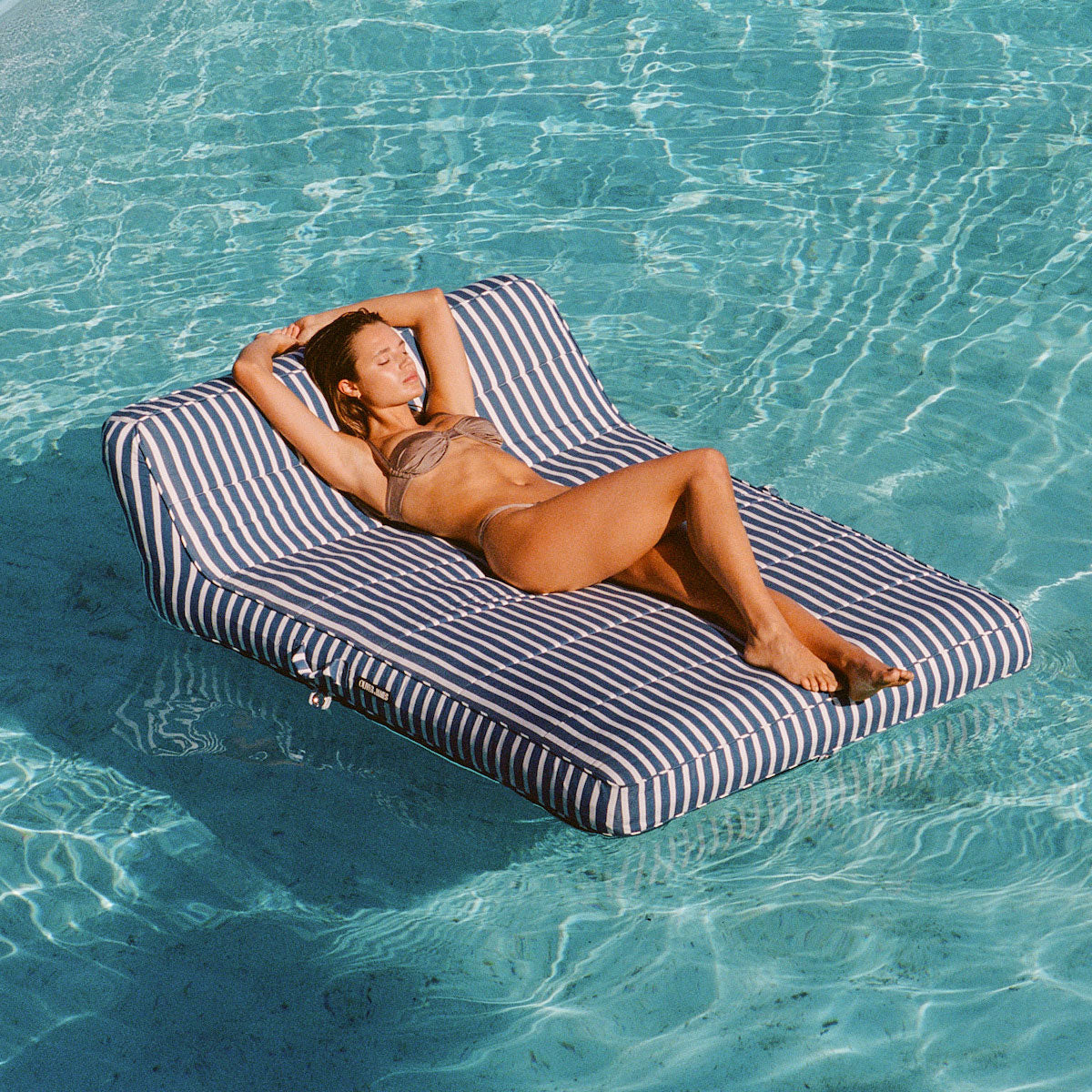 A women relaxing on a double white and blue striped pool float for adults in the middle of a swimming pool.