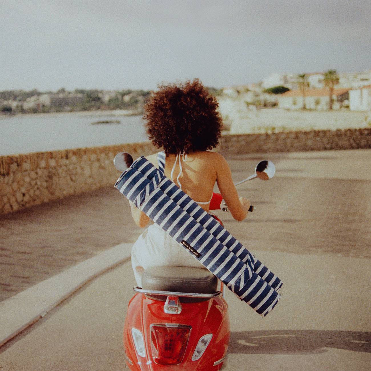 A women on a red moped with the sea in the background and a pool hammock for adults on her back.