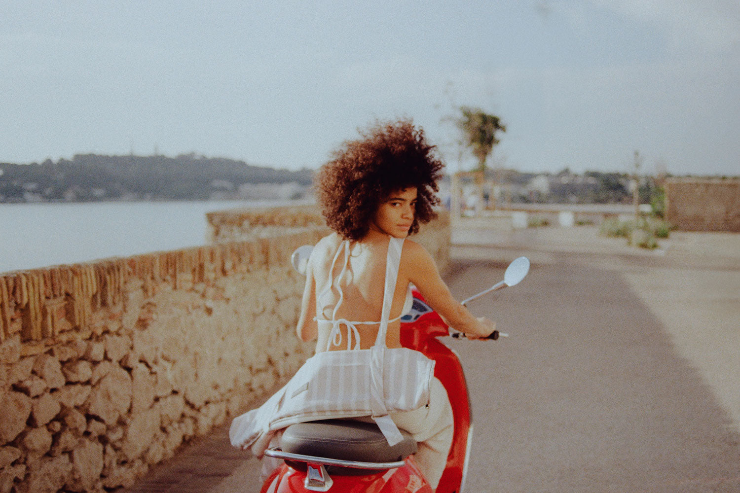 A women driving on a red vespa next to the sea with a deflated pool hammock for adults strapped to her back.