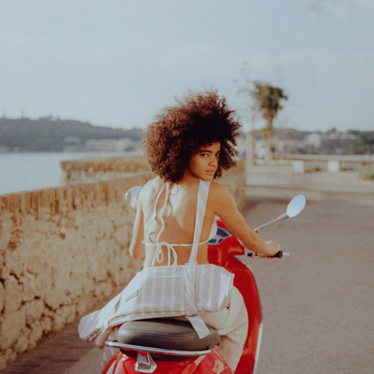A women on a red moped with the beach in the background and a pool toy for adults strapped to her back.