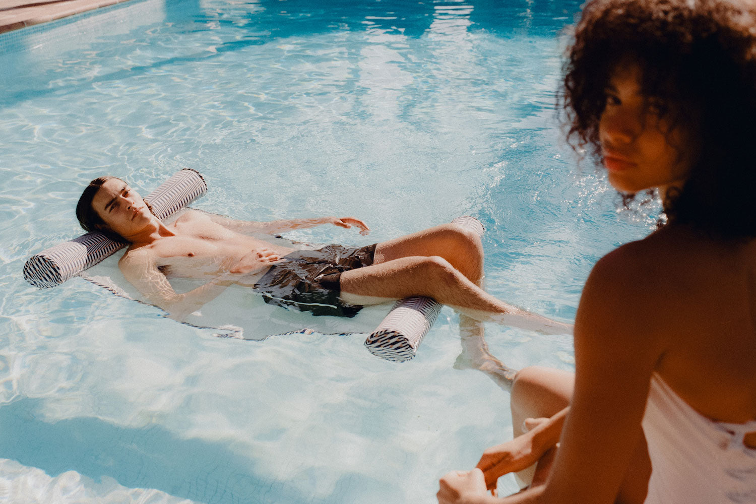 A young man floating on a luxury pool toy for adults in a swimming pool when a lady sitting in the foreground.